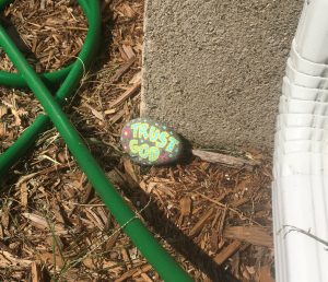 a rock that says, "Trust God," on the ground beside a garden hose